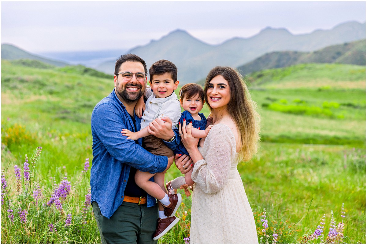 family of 4 smiling among wild flowers and green hills
